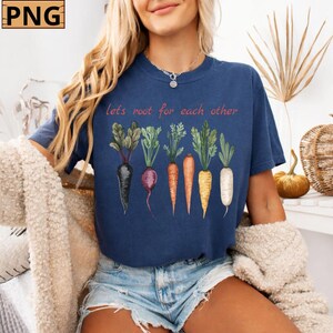 Puede incluir: Camiseta azul marino con una ilustración de acuarela de varias hortalizas de raíz, incluyendo zanahorias y remolachas. El texto "lets root for each other" está impreso encima de las verduras. La camiseta es de estilo casual.