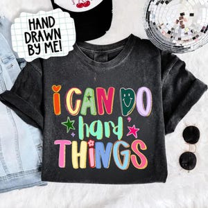 Pode incluir: T-shirt preto com a frase colorida "I CAN DO hard THINGS" em letras divertidas. O design inclui um coração, estrelas e uma carinha sorridente. Um balão de texto diz "HAND DRAWN BY ME!"