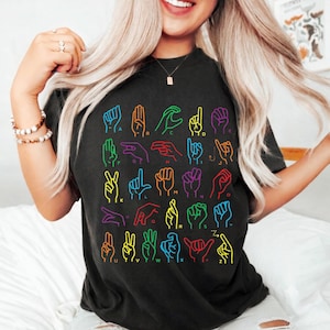 Könnte beinhalten: Ein schwarzes Kurzarm-T-Shirt mit einem bunten Grafikdruck des amerikanischen Gebärdensprachalphabets. Jeder Buchstabe von A bis Z wird durch eine neonfarbene Handzeichen-Illustration dargestellt, wobei der Buchstabe darunter gedruckt ist.