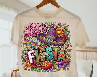 May include: Cream-colored t-shirt with a colorful "Let's Fiesta" graphic. The design includes a sombrero, flowers, a guitar, maracas, a chili pepper, and a piñata. The text is in various colors and fonts.