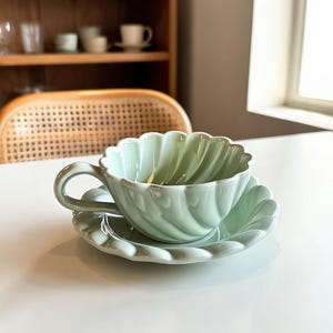 Könnte beinhalten: Ein hellgrünes Tee-Set, bestehend aus einer Tasse und einer Untertasse. Die Tasse und die Untertasse haben einen gewellten Rand. Die Tasse hat einen gebogenen Henkel. Das Set ist aus Keramik. Die Tasse steht auf der Untertasse auf einer weißen Oberfläche.