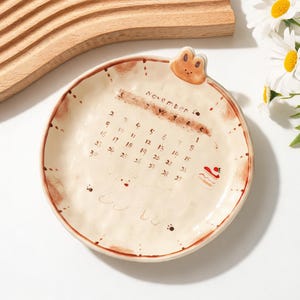 Puede incluir: Un plato redondo de cerámica con superficie color crema que presenta un calendario de noviembre en tonos marrones, una pequeña cabeza de oso marrón en el borde superior, un dibujo lineal de un perro y una ilustración de un pastel con fresa.