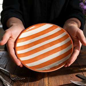 Może przedstawiać: Okrągły ceramiczny talerz z białym i pomarańczowym wzorem w paski. Talerz jest trzymany na tle ciemnego tła. Sztućce i wazon z fioletowymi kwiatami znajdują się na drewnianym stole.
