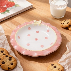May include: A pink and white polka dot ceramic plate with a scalloped edge and a yellow bow detail. Chocolate chip biscuits, a glass of milk, and a book with strawberry illustrations are also present.