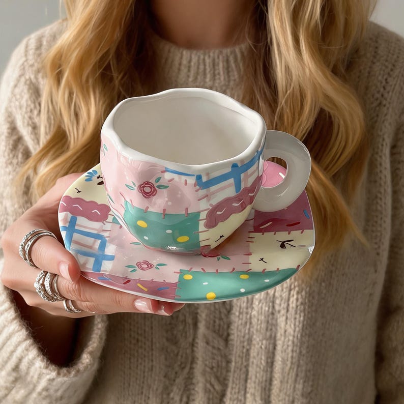 K&ouml;nnte beinhalten: Ein Keramik-Teebecher- und Untertassen-Set mit Patchwork-Design. Die Tasse und die Untertasse weisen eine Pastellfarbpalette mit rosa, blauen, gr&uuml;nen und gelben Quadraten sowie floralen Akzenten auf. Die Tasse hat einen wei&szlig;en Innenraum und einen abgerundeten Griff.