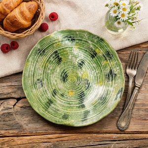 Könnte beinhalten: Ein grüner Keramikteller mit Spiralmuster, mit Grüntönen, Weiß und Schwarz. Der Teller steht auf einem Holztisch, mit einem Korb mit Croissants, Himbeeren, einer kleinen Vase mit Blumen, einer Gabel und einem Messer.