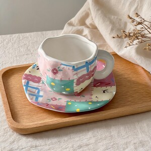 K&ouml;nnte beinhalten: Ein skurriles Keramik-Tee-Tassen- und Untertassen-Set. Die Tasse und die Untertasse weisen ein Patchwork-Design in Pastellrosa, Blau, Gr&uuml;n und Lila auf. Die Tasse hat einen unregelm&auml;&szlig;igen Rand und einen abgerundeten Griff. Das Set steht auf einem Holztablett.