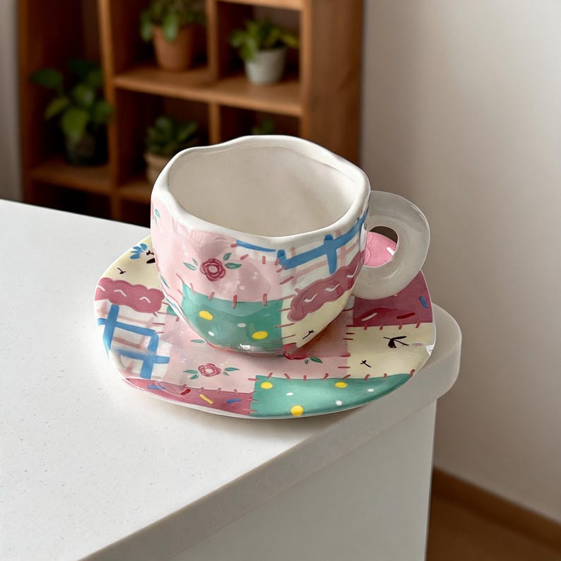 A 300ml hand-painted ceramic mug and saucer set displayed next to a matching 20cm dessert plate, all featuring a colorful patchwork quilt design.
