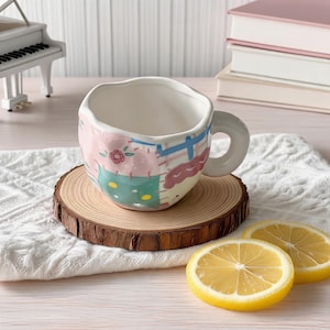 K&ouml;nnte beinhalten: Eine skurrile Keramiktasse mit einem Patchwork-Design in Pastellrosa, Gr&uuml;n und Blau. Die Tasse steht auf einem Holzuntersetzer mit nat&uuml;rlicher Kante. Zwei Zitronenscheiben befinden sich im Vordergrund. Im Hintergrund ist ein kleines wei&szlig;es Klavier zu sehen.