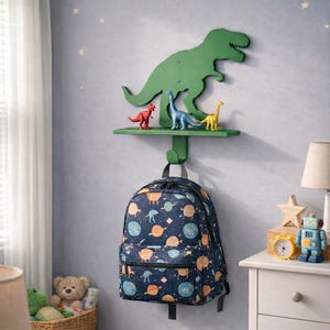 May include: A green dinosaur-shaped wall shelf with a hook, holding a backpack and small dinosaur figurines. The backpack is navy blue with a pattern of planets and stars. The room has a blue rug and a white dresser.