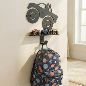 May include: A black metal wall shelf with a monster truck silhouette and a row of toy monster trucks. Below, a backpack with a space-themed pattern hangs from a hook. The shelf and truck are mounted on a white wall.