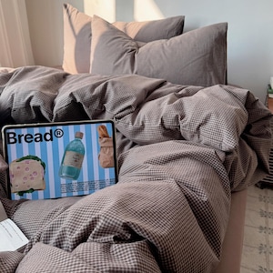 Könnte beinhalten: Ein Bett mit einer braun-weiß karierten Bettdecke und passenden Kissen. Ein Tablet mit dem Wort "Bread®" und Bildern von Lebensmitteln liegt auf dem Bett. Die Bettwäsche ist ordentlich arrangiert und schafft eine gemütliche Atmosphäre.