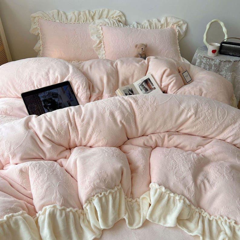 May include: A light pink bed set with a floral embossed pattern. The set includes a comforter, pillows, and a bedspread with ruffled edges. A tablet and a book are on the bed. A small teddy bear sits on the pillows.