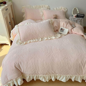 May include: A pink bed set with a floral pattern, including a comforter and pillows. The comforter and pillowcases have ruffled edges. A small teddy bear and a book are on the bed.