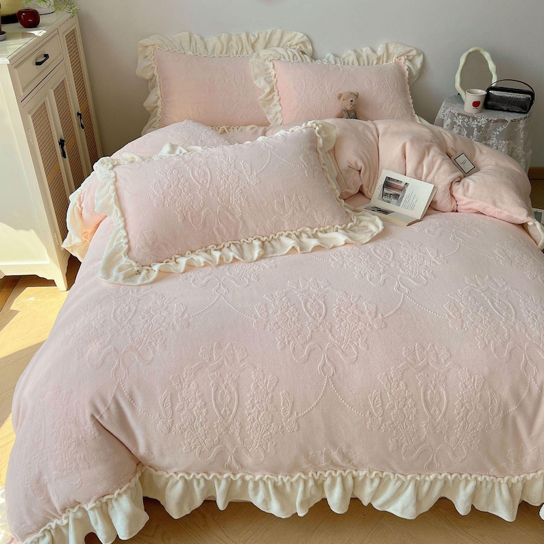 May include: A pink bed set with a floral pattern, including a comforter and pillows. The comforter and pillowcases have ruffled edges. A small teddy bear and a book are on the bed.