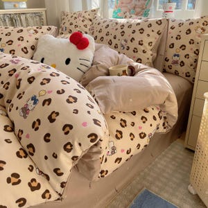 Könnte beinhalten: Ein Bett mit beiger Bettwäsche im Leopardenmuster, einschließlich Bettdecke und Kissenbezügen, sowie einem großen Hello Kitty-Kissen. Das Muster weist braune und schwarze Flecken, rosa Herzen und Hello Kitty-Illustrationen auf. Eine kleine Tasse ist zu sehen.