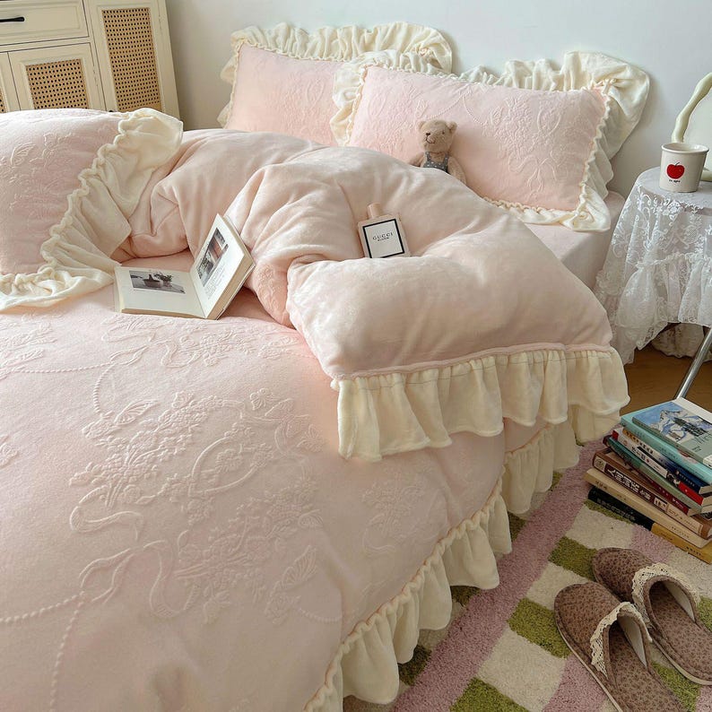 May include: A soft pink bedding set with cream-colored ruffled edges. The set includes a comforter, pillows, and a fitted sheet with an embossed floral pattern. A small teddy bear and a book are on the bed.
