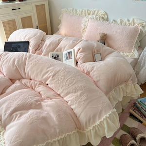 May include: A soft pink bedding set with a floral embossed pattern. The set includes a comforter, two pillows with ruffled edges, and a bed skirt. A laptop and a book are visible on the bed, along with a small teddy bear.