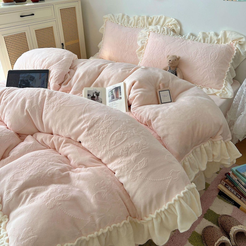 May include: A soft pink bedding set with a floral embossed pattern. The set includes a comforter, two pillows with ruffled edges, and a bed skirt. A laptop and a book are visible on the bed, along with a small teddy bear.