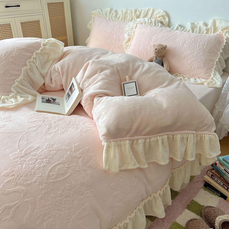 May include: A light pink bedspread with a floral pattern and cream-colored ruffled trim. The bed is made with a plush, light pink comforter and matching pillowcases. A small teddy bear and a perfume bottle are on the bed.
