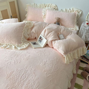 May include: A light pink bedding set with floral embossed patterns. The set includes a comforter, pillows, and pillowcases, all trimmed with cream-colored ruffles. A book and perfume bottle rest on the comforter. A small teddy bear sits on the pillows.