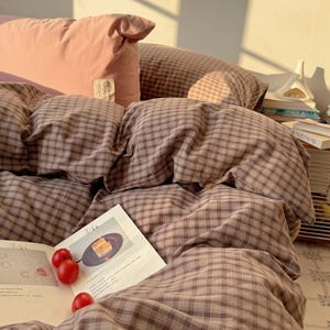 May include: A bed with a brown and white checked duvet cover and pillowcases. A pink bolster pillow is at the head of the bed. A magazine with a photo of a cake and red cherries is on the bed.