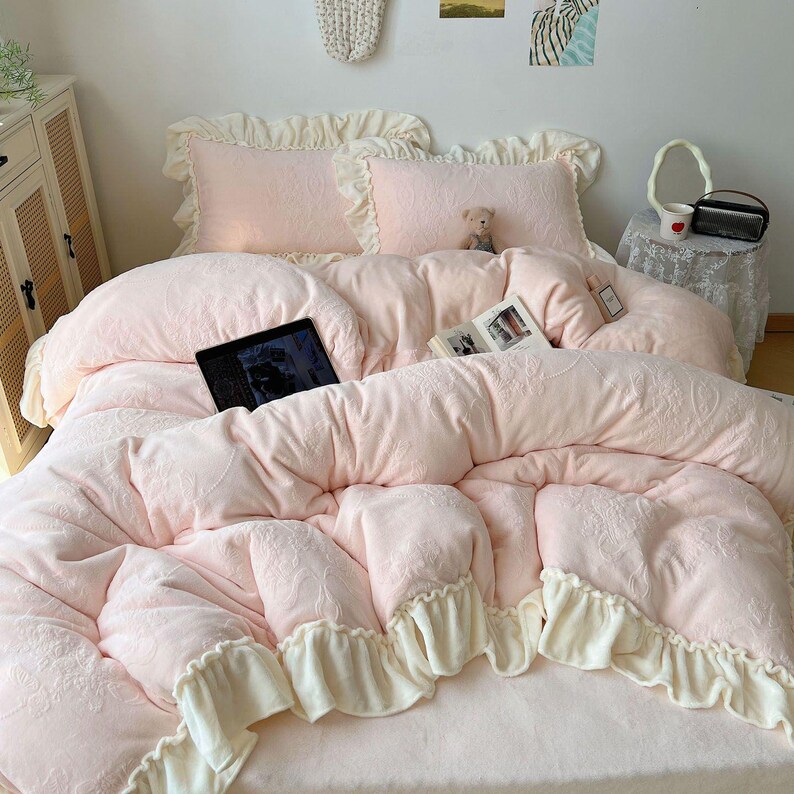 May include: A soft pink bedding set with a floral pattern, featuring a comforter, pillows, and a bed sheet. The comforter has a ruffled edge in a cream color. A laptop and a book are on the bed. A small teddy bear is on the pillows.
