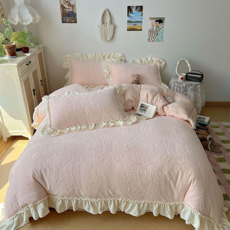 May include: A light pink bedding set with a floral pattern, featuring ruffled edges and pillowcases. The set includes a comforter and pillowcases, creating a soft and inviting aesthetic. A small teddy bear sits on the bed.
