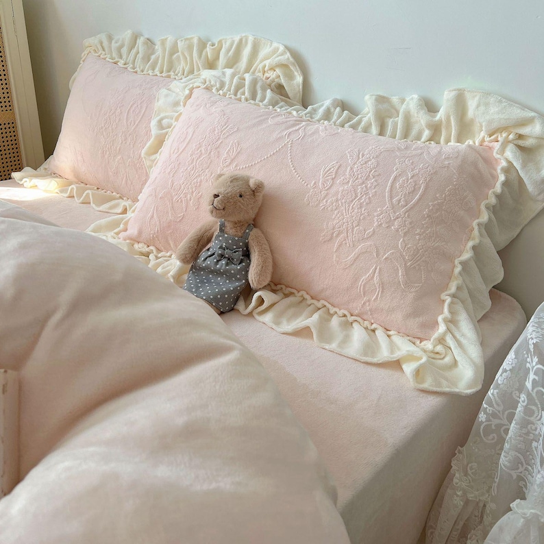 May include: A bed with pink bedding, including two decorative pillows with floral embroidery and cream-colored ruffled edges. A small teddy bear in a gray polka-dot dress sits on the bed. The bedding has a soft, inviting appearance.