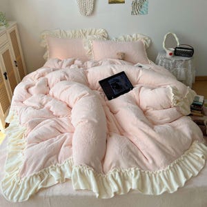 May include: A light pink bed set with a ruffled edge. The set includes a comforter, two pillows, and a bedspread. The comforter and pillows have a floral pattern. A tablet is on the bed. The bed set is made of a soft material.