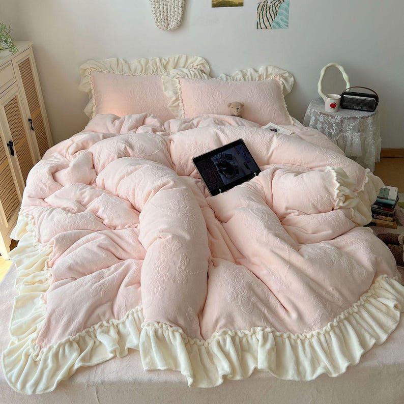 May include: A light pink bed set with a ruffled edge. The set includes a comforter, two pillows, and a bedspread. The comforter and pillows have a floral pattern. A tablet is on the bed. The bed set is made of a soft material.