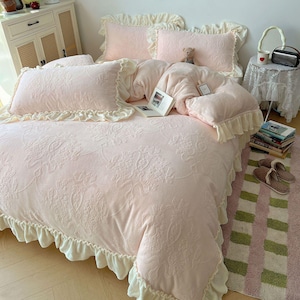 May include: A light pink bed set with a floral pattern, ruffled edges, and matching pillowcases. The bedding includes a comforter and pillows. A small teddy bear and an open book are on the bed. The bed set is on a pink and green striped rug.