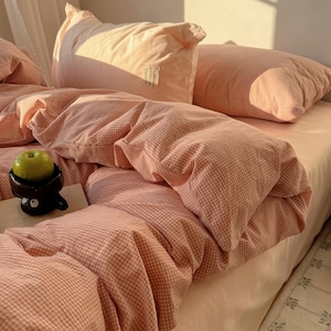 Könnte beinhalten: Ein Bett mit einem hellpfirsichfarbenen Spannbettlaken, Kissen und einem Bettbezug. Der Bettbezug und die Kissen haben ein kleines, hellrosa und weiß kariertes Muster. Ein Apfel liegt in einer kleinen schwarzen Tasse auf dem Bett.