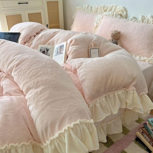 May include: A light pink bed set with a floral pattern, featuring a comforter, pillows, and a bolster pillow. The comforter and pillows have ruffled edges. A book and a perfume bottle are on the bed. A small teddy bear is on the pillow.
