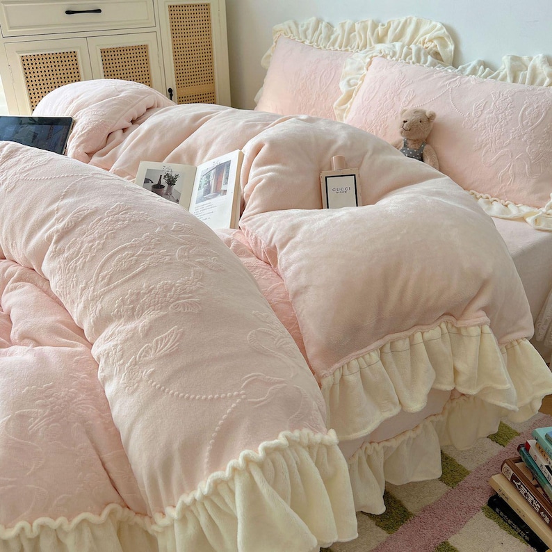 May include: A light pink bed set with a floral pattern, featuring a comforter, pillows, and a bolster pillow. The comforter and pillows have ruffled edges. A book and a perfume bottle are on the bed. A small teddy bear is on the pillow.