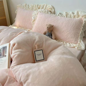 May include: A cozy bed setting with light pink bedding, including a plush duvet and embroidered pillows with ruffled edges. A small teddy bear sits on the bed, and a Gucci Bloom perfume bottle rests on the duvet. A book is also visible.