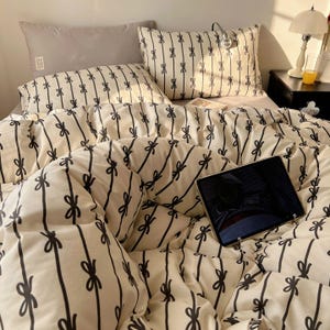 Może przedstawiać: Ł&oacute;żko z kremową kołdrą i poszewkami na poduszki z czarnym wzorem kokardki. Widoczna jest r&oacute;wnież szara poduszka i tablet. Mała lampa i stolik nocny znajdują się w tle.
