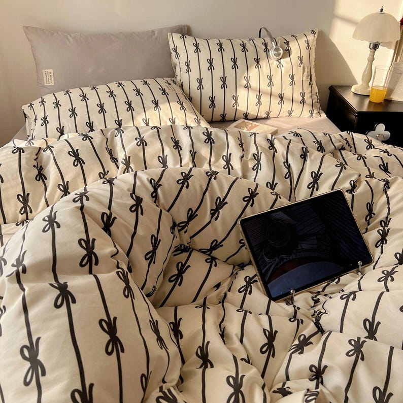 Może przedstawiać: Ł&oacute;żko z kremową kołdrą i poszewkami na poduszki z czarnym wzorem kokardki. Widoczna jest r&oacute;wnież szara poduszka i tablet. Mała lampa i stolik nocny znajdują się w tle.