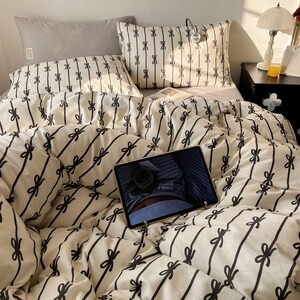 Może przedstawiać: Kremowy zestaw pościeli z czarnym wzorem kokardek. Zestaw zawiera kołdrę, poduszki i szarą poduszkę. Tablet jest umieszczony na ł&oacute;żku. Pok&oacute;j jest oświetlony lampką nocną.