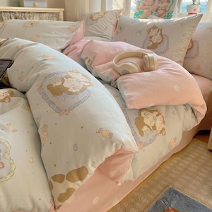 May include: A light blue and pink bedding set with a cartoon animal pattern. The set includes a duvet cover, pillows, and a fitted sheet. A pair of white headphones rests on the bed. The bedding has a soft, inviting appearance.