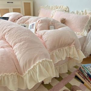 May include: A soft pink bedspread with a floral embossed pattern and a cream-colored ruffled edge. The bedding includes matching pillows and a long bolster pillow. A small teddy bear sits on the bed.