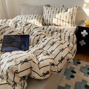 Może przedstawiać: Ł&oacute;żko z kremową kołdrą i poszewkami na poduszki z czarnym wzorem kokardek i pask&oacute;w. Szara poduszka i tablet r&oacute;wnież znajdują się na ł&oacute;żku. Ł&oacute;żko jest pościelone i gotowe do użytku.