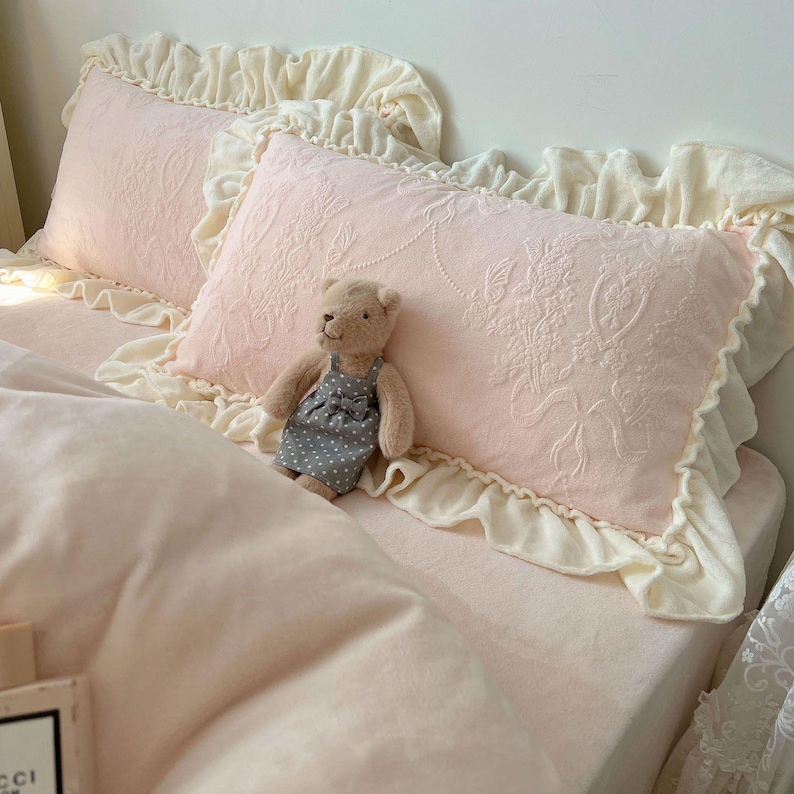 May include: Two pink rectangular pillows with floral embroidery and cream-colored ruffled edges rest on a bed. A small teddy bear wearing a gray polka-dot dress sits on the bed. The bedding is a soft pink color.