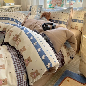 Puede incluir: Una cama cubierta con un juego de ropa de cama beige y blanco con temática de ositos de peluche. El juego incluye una funda nórdica, almohadas y una sábana bajera. Las almohadas tienen un borde azul con ositos de peluche blancos y corazones. Un osito de peluche marrón y auriculares negros están en la cama.