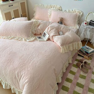 May include: A light pink bed set with a floral pattern, ruffled edges, and matching pillows. A teddy bear and a book are on the bed. The bed set is on a pink and green striped rug.
