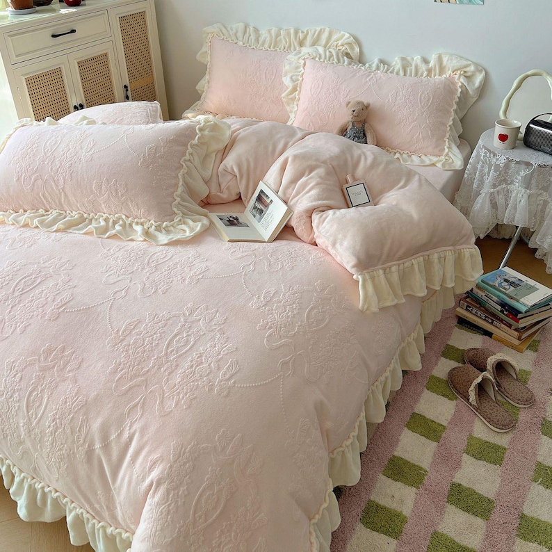 May include: A light pink bed set with a floral pattern, ruffled edges, and matching pillows. A teddy bear and a book are on the bed. The bed set is on a pink and green striped rug.
