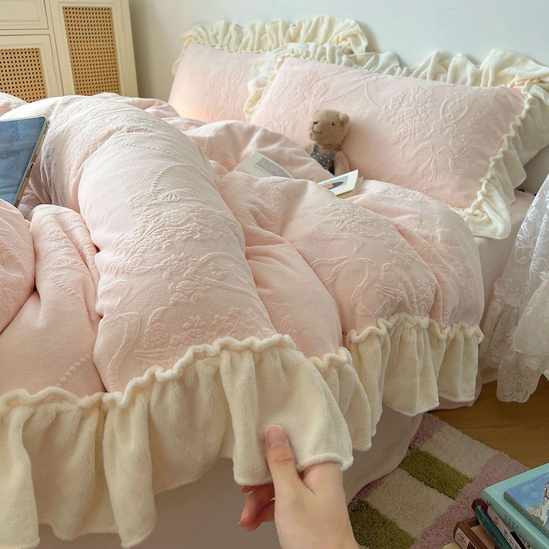 May include: A bed with a light pink comforter and matching pillowcases. The comforter and pillowcases have a floral pattern and are trimmed with a cream-colored ruffle. A small teddy bear sits on the bed.