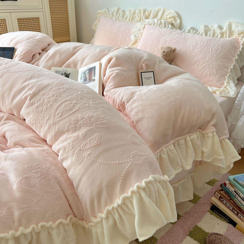 May include: A soft, pink bedding set with a floral embossed design. The set includes a comforter, pillows, and a bed skirt with ruffled edges. A book and a perfume bottle are on the bed. A small teddy bear is nestled among the pillows.