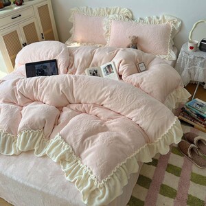 May include: A soft pink bed set with a floral pattern, featuring a comforter, pillows, and ruffled edges. A laptop and books are on the bed. The bed set is made of a plush material, creating a cozy and inviting atmosphere.