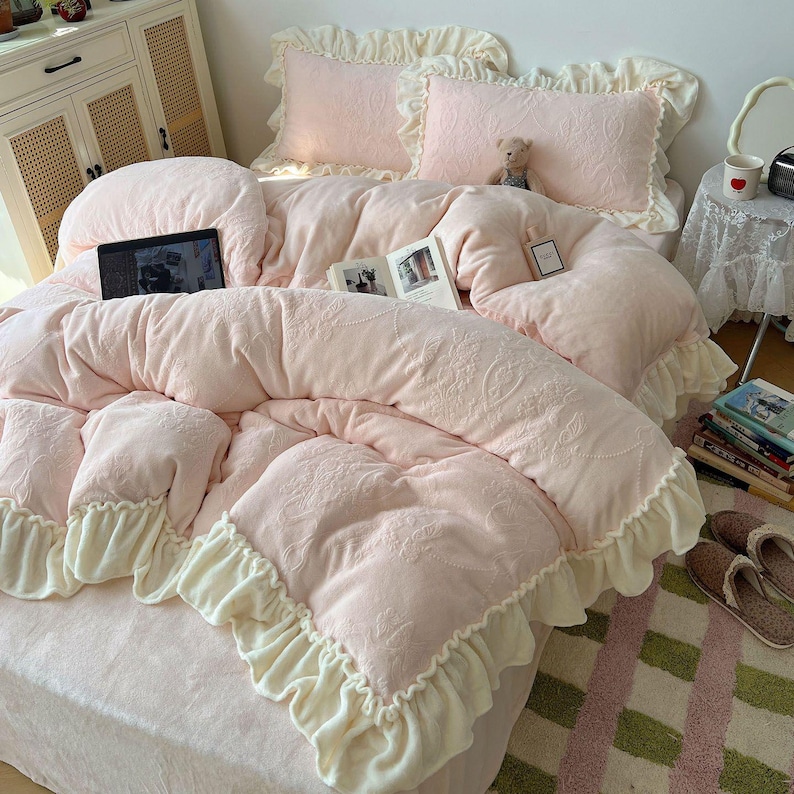 May include: A soft pink bed set with a floral pattern, featuring a comforter, pillows, and ruffled edges. A laptop and books are on the bed. The bed set is made of a plush material, creating a cozy and inviting atmosphere.